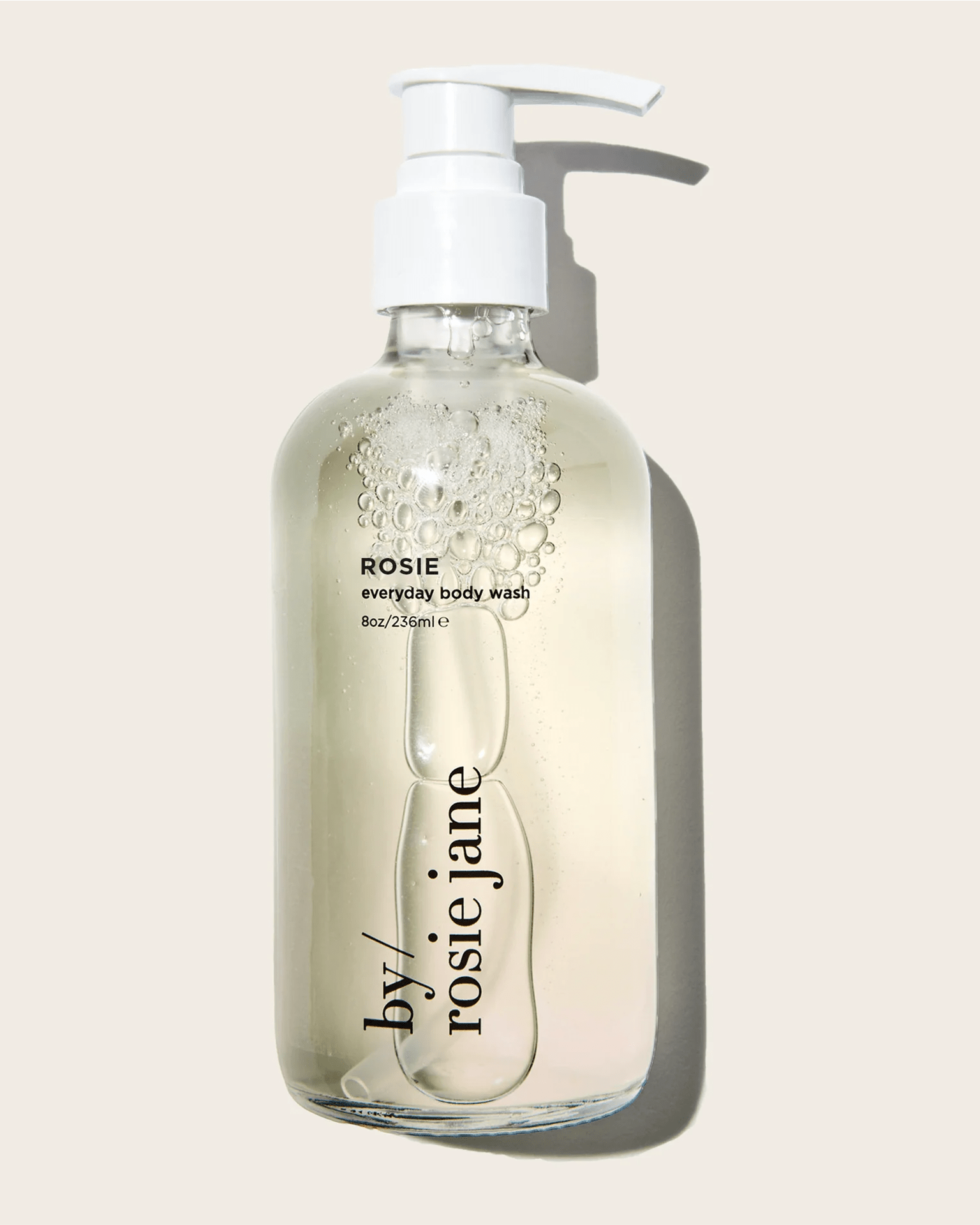 A clear pump bottle labeled Rosie Everyday Body Wash by Rosie Jane, filled with transparent liquid and bubbles, sits against a light beige background—featuring a gentle white musk scent.