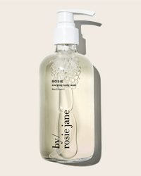 A clear pump bottle labeled Rosie Everyday Body Wash by Rosie Jane, filled with transparent liquid and bubbles, sits against a light beige background—featuring a gentle white musk scent.