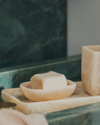 A bar of soap rests on the Saarde Flow Resin Soap Dish in Peach Blush, which sits on a marble tray beside a marble cup, all arranged on a green marble countertop.