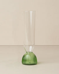 The Saarde Kairos Champagne Flute in Green, featuring a tall, clear design with a textured green base, is set upright on a light-colored surface against a plain background.