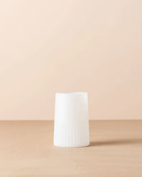 The Saarde Kairos Large Water Glass in Opaque White, featuring a cylindrical shape with a ridged lower half, stands upright on a beige surface against a light beige background.