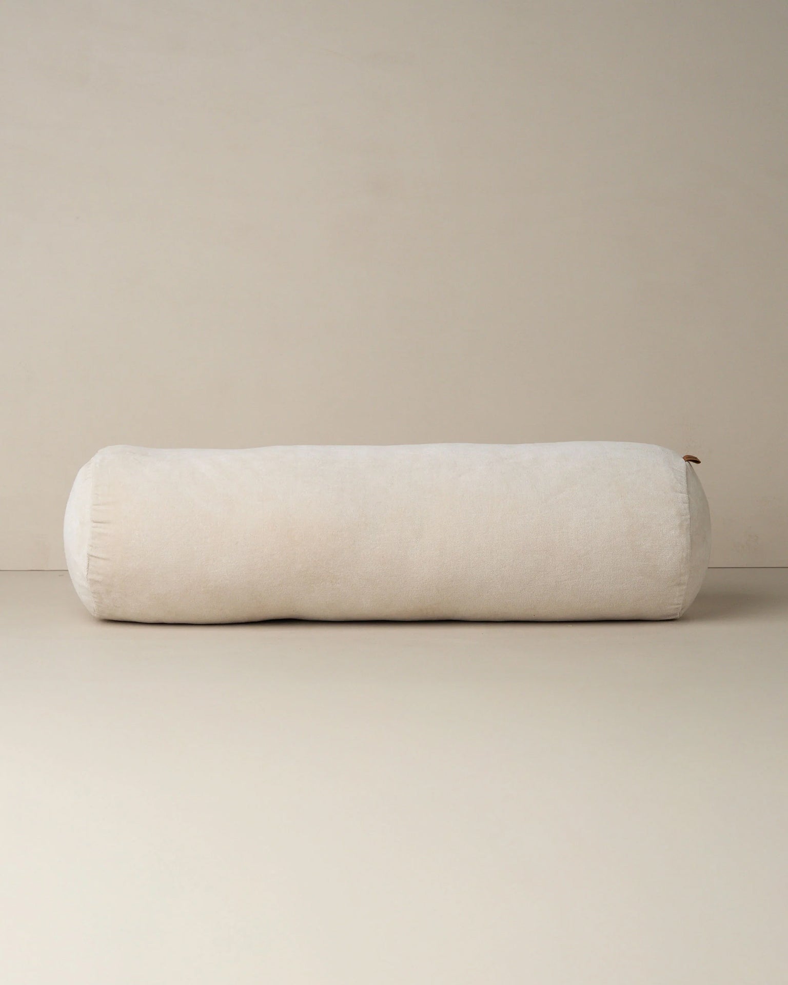 The Saarde Velvet Structured Bolster in Clay, featuring a large cylindrical shape, rests on a neutral beige surface with a matching backdrop.