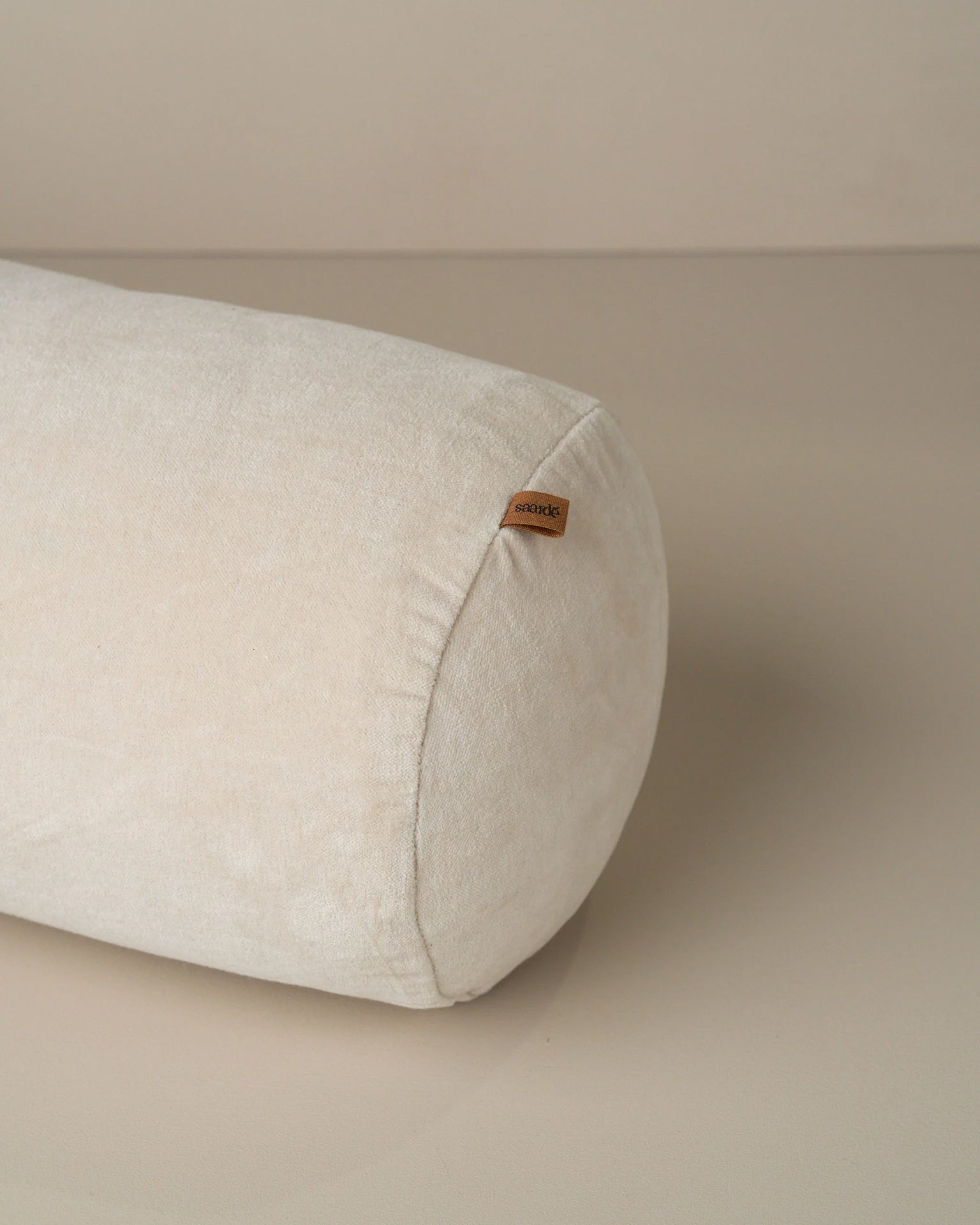The Saarde Velvet Structured Bolster in Clay is a cylindrical off-white cushion with a small brown fabric tag on one end, displayed on a smooth, neutral surface.