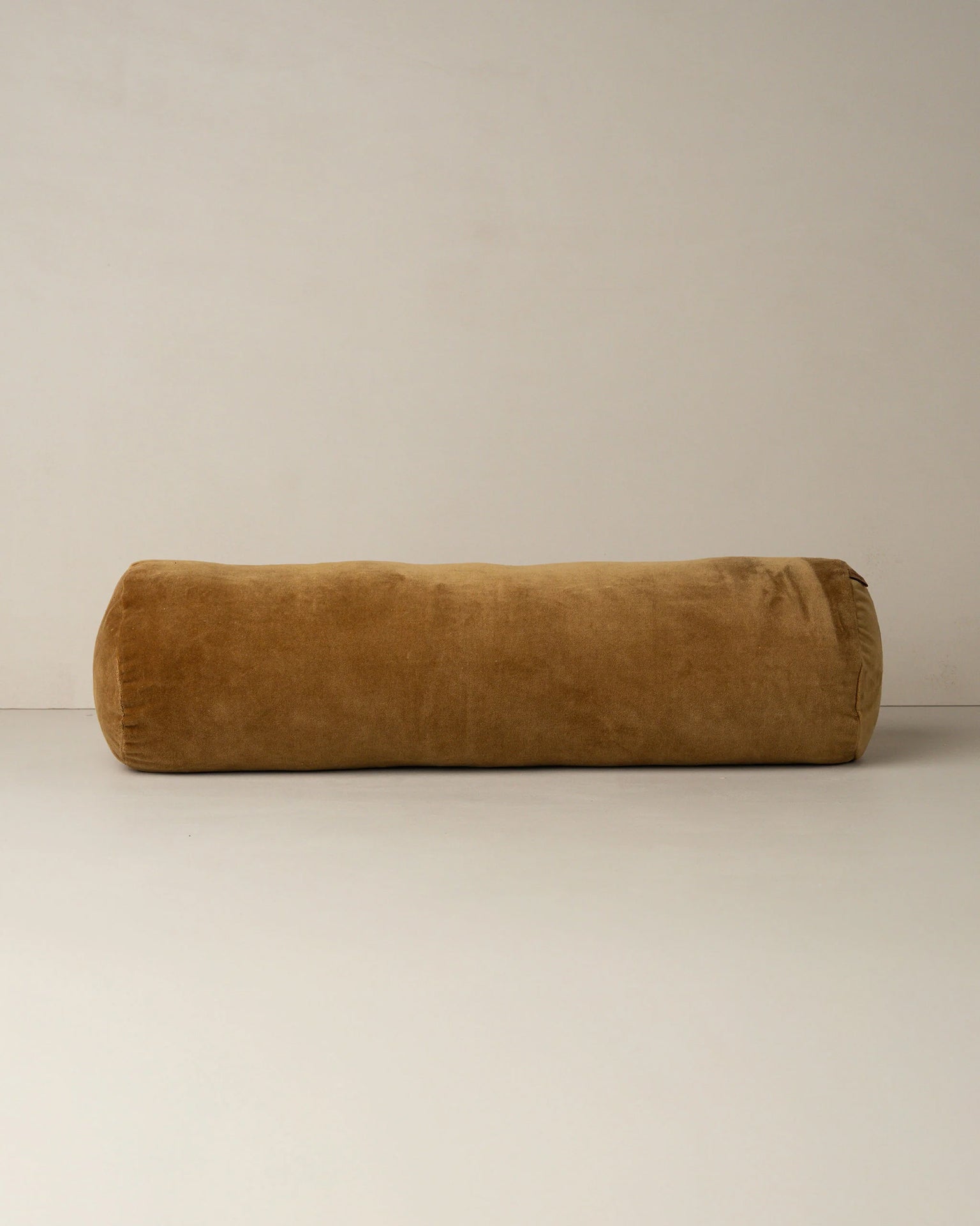 The Saarde Velvet Structured Bolster in Nutmeg, a long, cylindrical velvet cushion in rich brown, rests on a plain light surface against a neutral wall.