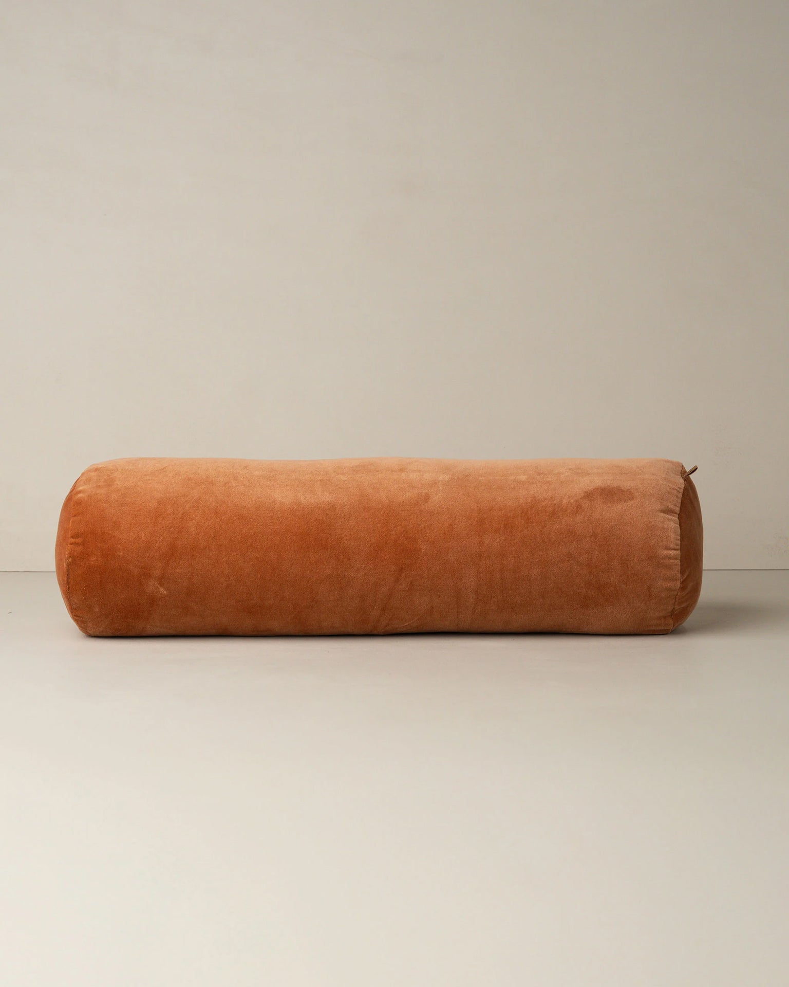 The Saarde Velvet Structured Bolster in Terracotta rests on a neutral floor beside a plain wall, showcasing its large cylindrical cotton velvet design.