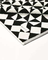 The Slowtide Gatsby Bath Mat in Black, made from sustainable cotton, showcases a geometric triangle design and a visible fabric tag, displayed on a white background.