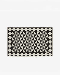 The Gatsby Bath Mat in Black by Slowtide features a black and white geometric pattern, is made from sustainable cotton, and has a small corner label.