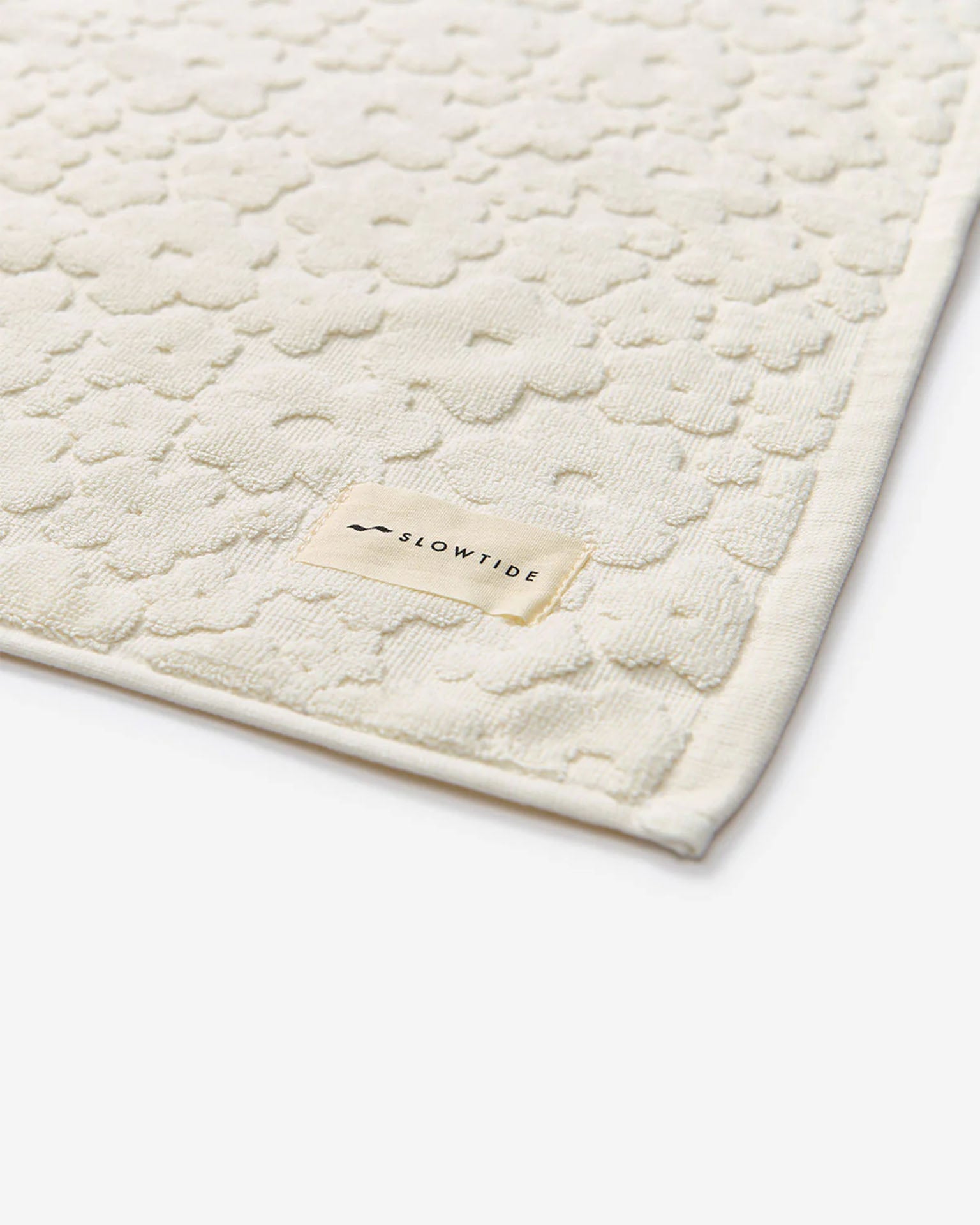 A close-up of the Ginny Bath Mat by Slowtide, a cream-colored, absorbent mat made from sustainably sourced cotton with a textured floral pattern and a branded label stitched on the edge.