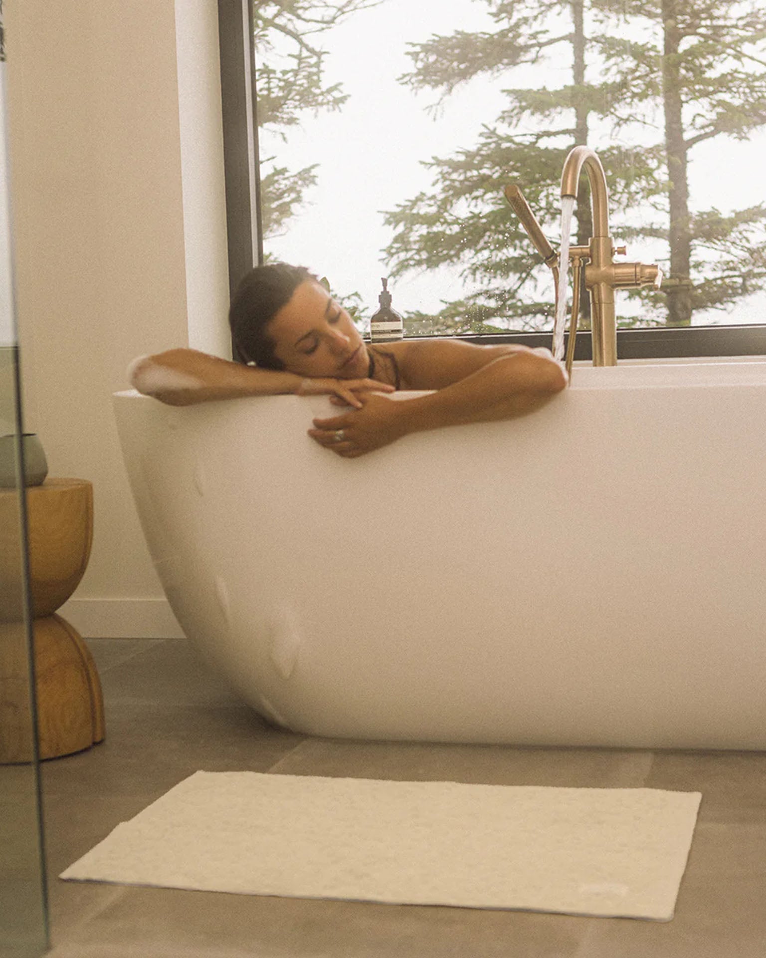 A person relaxes in a white bathtub near a window with trees outside; the Ginny Bath Mat by Slowtide, made from sustainably sourced cotton, lies on the floor, while a wooden table sits beside the tub.