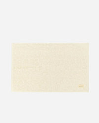 The Ginny Bath Mat by Slowtide is a rectangular, cream-colored mat made from absorbent, sustainably sourced cotton with a subtle floral pattern and a small label in the bottom right corner, shown against a plain background.