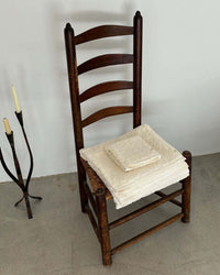 A wooden ladder-back chair with folded Slowtide Ginny Bath Towels on the seat sits on a concrete floor beside a metal candle holder with two candles.