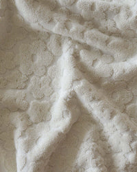 The Ginny Bath Towel by Slowtide features an off-white, textured fabric with a raised floral or hexagonal pattern, crafted from sustainably sourced cotton for plush, spa-like comfort and eco-friendly softness.