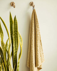 The Slowtide Ginny Bath Towel (600 GSM), with its yellow pattern, hangs beside a tall snake plant on a light wall, adding spa-like comfort and style to your space.