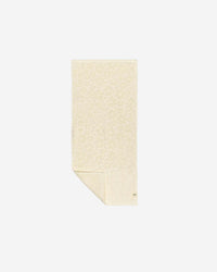 The Slowtide Ginny Hand Towel, in light beige with a subtle floral pattern, is displayed flat on a white background with one corner gently folded.