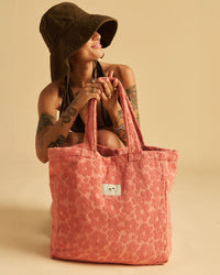 A person kneels against a beige background, holding the Slowtide Ginny Terry Tote Bag in Guava—crafted from sustainably sourced cotton and featuring a white label—while wearing a brown bucket hat and dark bikini top.