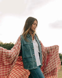 A woman stands outdoors with arms extended, draped in the Slowtide Palaka L/W Beach Blanket in Guava over a denim jacket, white shirt, and jeans.