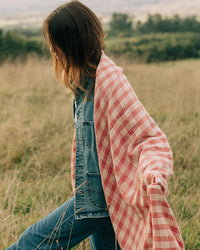 A person in blue denim and a pink checkered shawl carries the Slowtide Palaka L/W Beach Blanket in Guava across a grassy field, with trees and hills in the background.
