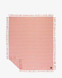 The Slowtide Palaka L/W Beach Blanket in Guava features a rectangular pink and white checkered design with fringed edges, shown flat with one corner slightly folded.
