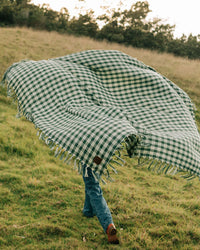 A person walks on grass holding the Slowtide Palaka L/W Beach Blanket in Sequoia, a large green and white checkered blanket that billows and covers most of their body.