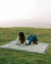 A person lies on their side on the Slowtide Palaka L/W Beach Blanket in Sequoia, spread across grass, gazing away toward a misty landscape.