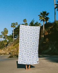On a sandy beach under a clear sky, only their feet show as someone holds up Slowtide’s The Dream Oversized Premium Woven Towel in Lagoon, framed by palm trees and lush greenery.