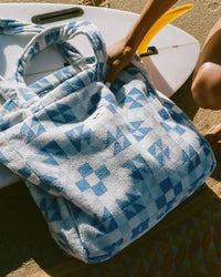 A person sets the Slowtide Dream Terry Tote Bag in Lagoon, crafted from sustainably sourced cotton, on a surfboard at the beach.