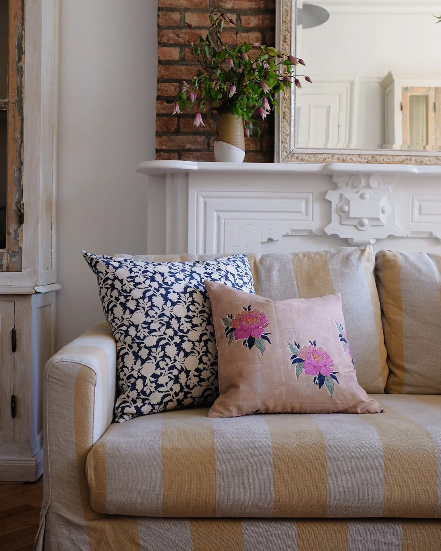 A beige striped sofa with two decorative pillows, including the Soil to Studio Babita Linen Pillow in Old Rose (18x18), sits before a white mantle and exposed brick wall.