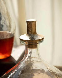 A close-up of The Collective’s Pebbled Decanter - Wide, featuring a hammered aluminum stopper, with another vessel of brown liquid blurred in the background.