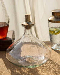 The Collectives Pebbled Decanter - Wide features a handcrafted glass body with a hammered aluminum stopper, shown on burlap with two additional glass containers in the background.