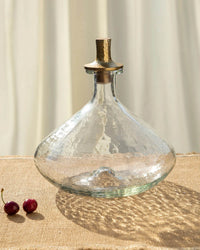 The Collective’s Pebbled Decanter - Wide features a handcrafted glass body with a brass handle, shown on a textured surface beside two cherries and a sheer curtain.
