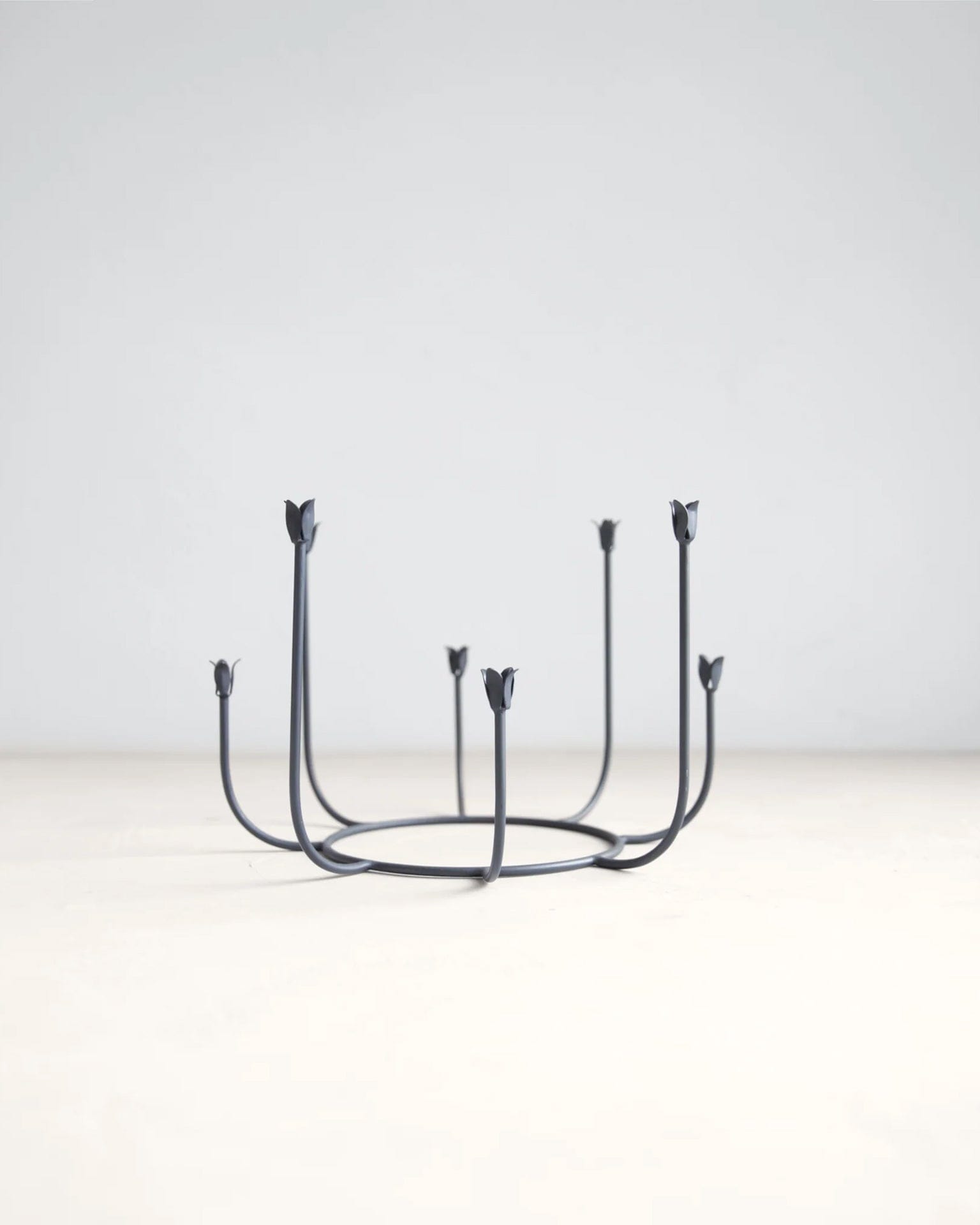 The Floral Societys Beacon Long Stem Candelabra in Smoke features six upward-curving arms with flower-shaped holders, crafted from minimalist black wrought iron and displayed on a light surface against a plain background.