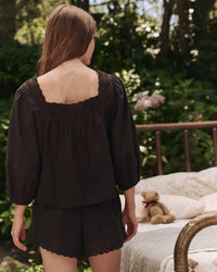 A woman wearing the Greats Eyelet Button Sleep Top in Black stands outdoors near a bed with a stuffed teddy bear, surrounded by greenery.
