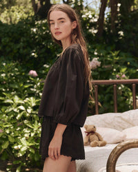 A woman with long brown hair stands outdoors in front of greenery, wearing the Great’s Eyelet Button Sleep Top in Black. A bed with a teddy bear is visible in the background.