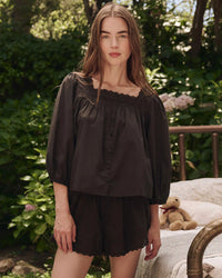 A woman stands outdoors before lush greenery, wearing the Great’s Eyelet Button Sleep Top in Black. A teddy bear rests on a bench beside her, adding a charming touch to the breezy setting.