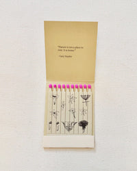 The Quiet Botanists Botanical Matches are an open floral matchbook with pink-tipped, biodegradable matches, each featuring unique black-and-white flower art and a Gary Snyder nature quote inside the cover.
