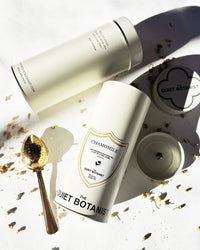Two cylindrical containers of The Quiet Botanist Chamomile Tea rest on a white surface with a gold tea strainer spoon and chamomile flowers, evoking a calming, caffeine-free herbal infusion.