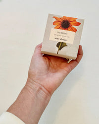 A hand holds a box labeled EVERYDAY - HANDMADE SOAP by The Quiet Botanist, featuring an orange flower design—ideal for sensitive skin and subtly fragranced with natural ingredients.