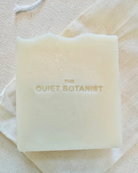 The Quiet Botanists Everyday - Handmade Soap is a white rectangular olive oil bar, embossed with the brand name and crafted for sensitive skin, featuring a subtle natural fragrance. Shown on a light fabric surface.