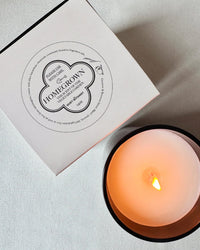 A lit Home Grown Candle by The Quiet Botanist made from natural coconut wax sits next to its white box on a white surface, filling the air with a subtle tomato leaf scent.