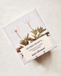 A box labeled Home Grown Candle by The Quiet Botanist, featuring tomato leaf, herbs & bergamot scents. Made with natural coconut wax and decorated with botanical illustrations on a light background.