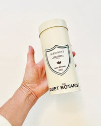 A hand holds a cylindrical tin of Iced Mint Tea by The Quiet Botanist against a plain white background, highlighting this refreshing, caffeine-free blend.