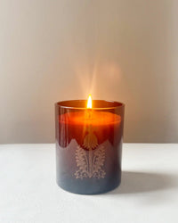 The Quiet Botanists In The Weeds candle, hand-poured into a brown glass holder with a decorative pattern, is displayed on a white surface against a plain background.