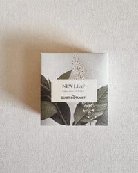 A square box labeled New Leaf (Nettle) - Handmade Soap by The Quiet Botanist, featuring a botanical illustration, rests on a textured surface, offering natural hydration and gentle care for skin and hair.