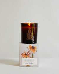 A lit brown glass Orchard Sun Candle by The Quiet Botanist rests on a white box with yellow flowers. Made from natural coconut wax and hand-poured, it adds a gentle glow to any space.