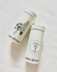 Two cylindrical containers from The Quiet Botanist rest on a light surface—one features a lilac flower design, while the other is labeled “Edible Flower Blend,” containing an Organic Edible Flower Blend that’s caffeine free.