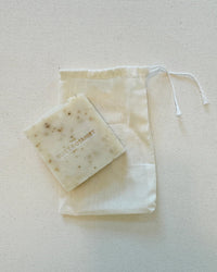 A square bar of Smell The Roses - Handmade Soap by The Quiet Botanist sits atop a white drawstring cloth bag, displayed on a light background.