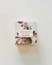 A small box labeled Smell The Roses - Handmade Soap by The Quiet Botanist, decorated with pink wild roses and green leaves on a light background, contains natural soap.