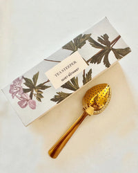 A gold, leaf-shaped tea steeper by The Quiet Botanist sits beside a floral box labeled “Tea Steeper”—an ideal gift for tea lovers.