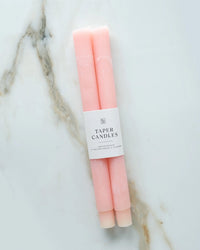 Two Coral Pink Taper Candles by BOTANICA, hand-dyed with a white label, are displayed on a white marble surface featuring light brown and gray veining.