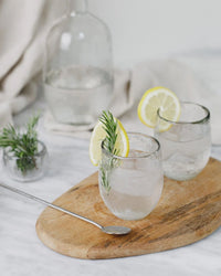 Two Pebbled Spirits Glasses by The Collective, filled with iced water and garnished with lemon and rosemary, sit on a wooden board. A carafe and a bowl of rosemary are visible in the background. Set of 4.